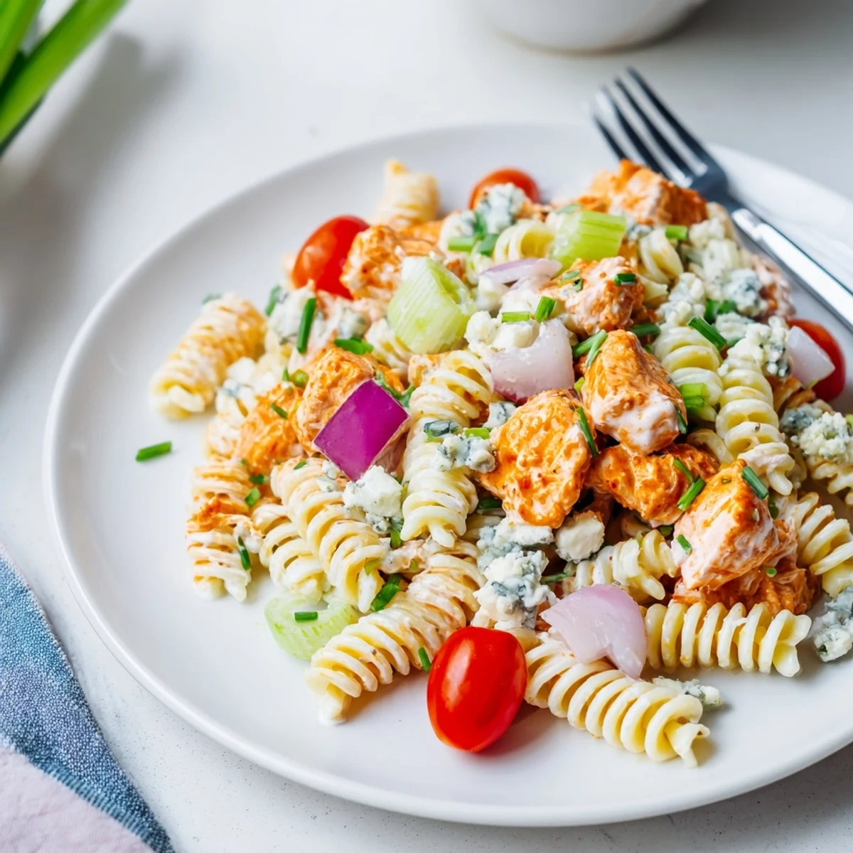 Bright red buffalo chicken pasta salad with crunchy celery, diced red bell peppers, and creamy ranch dressing garnished with crumbled blue cheese on a white plate.  